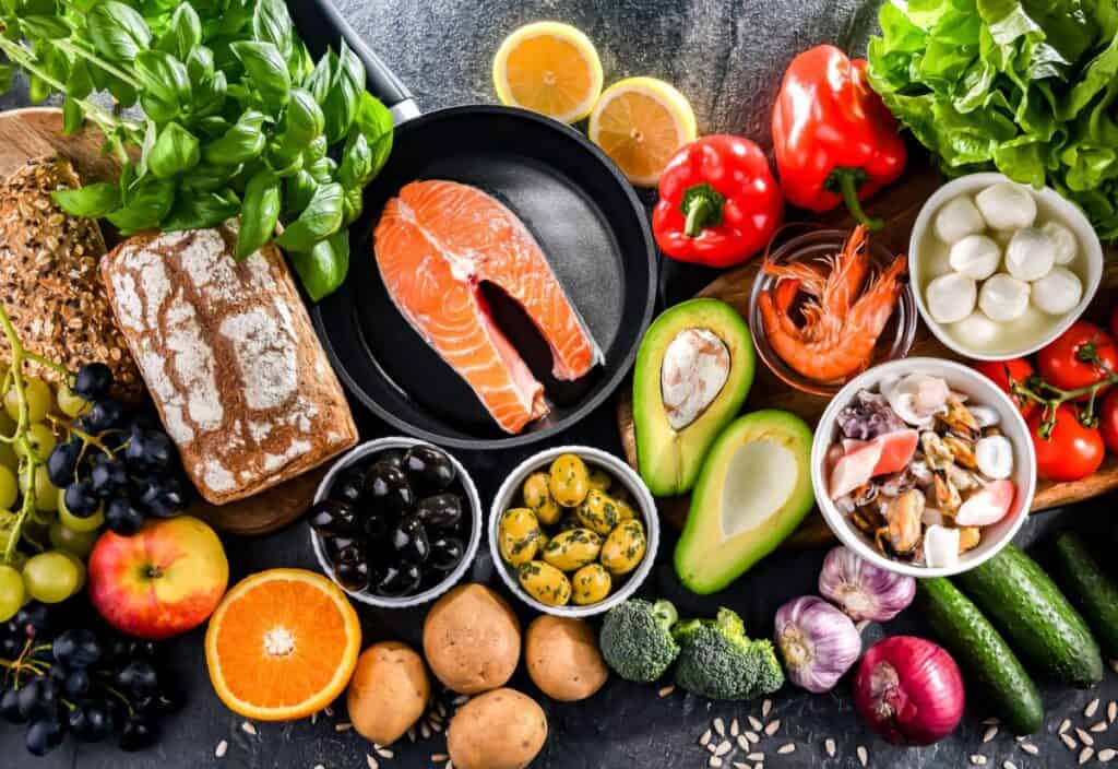 An assortment of fresh foods including salmon, bread, fruits, vegetables, olives, cheese, and seafood arranged on a dark surface.