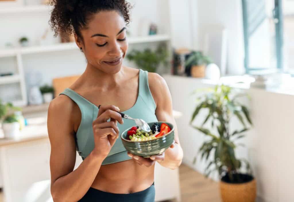 Woman in athletic wear holds a bowl of salad and eats with a fork in a bright, modern kitchen.