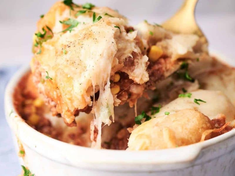 Close-up of a cowboy casserole in a white dish, showing layers of ground meat and vegetables topped with a golden-brown mashed potato crust, garnished with chopped parsley.
