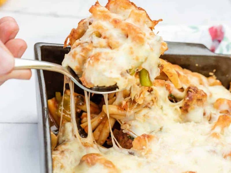A hand lifts a serving of meatball casserole from a rectangular baking dish, showing melted cheese stretching.
