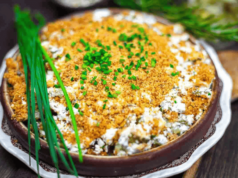 A casserole topped with golden breadcrumbs and chopped chives, served in a round brown dish, with fresh chives on the side.