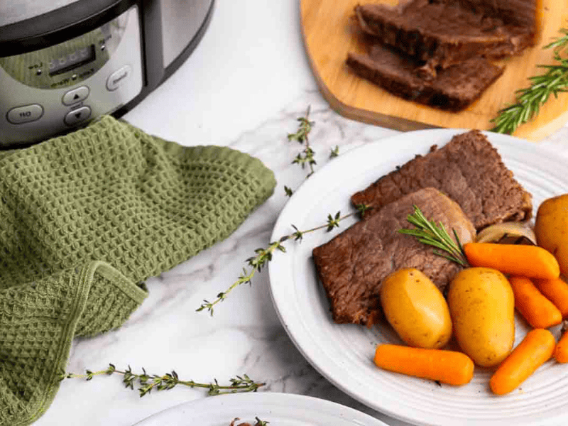 A plate with sliced pot roast, potatoes, and carrots sits on a marble surface next to a slow cooker, a green towel, and fresh herbs.